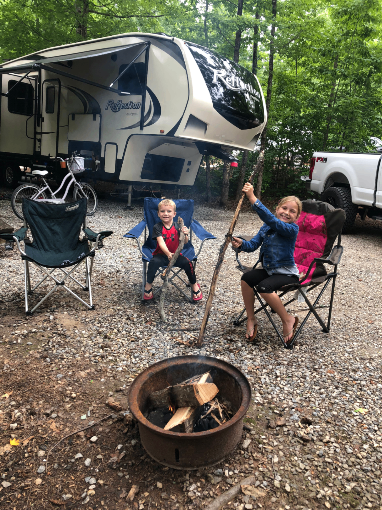 Two children roasting sticks over a campfire at a wooded campsite, with a Grand Design Reflection RV and pickup truck in the background