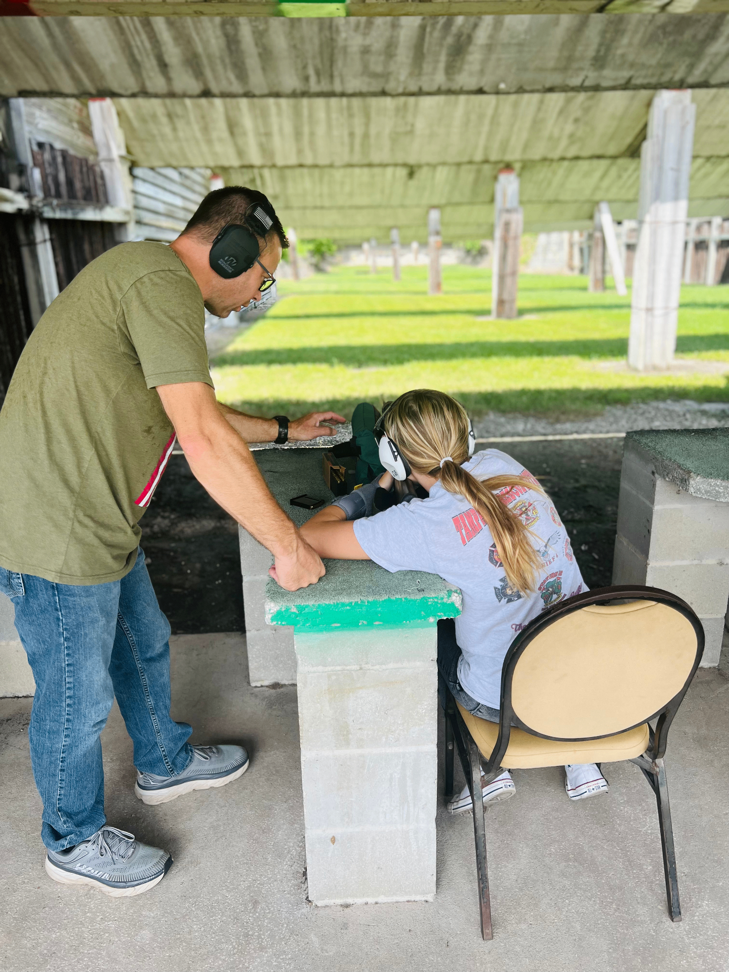 An adult man wearing ear protection guides a young girl with a ponytail as she learns to shoot at a covered outdoor rifle range
