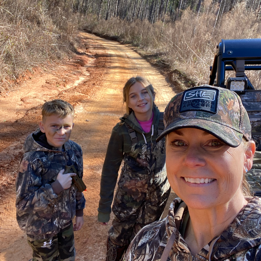 A woman and two children in camouflage smiling on a dirt trail with an off-road vehicle behind them