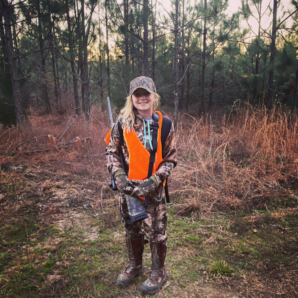 Child in camouflage and an orange vest standing in a pine forest