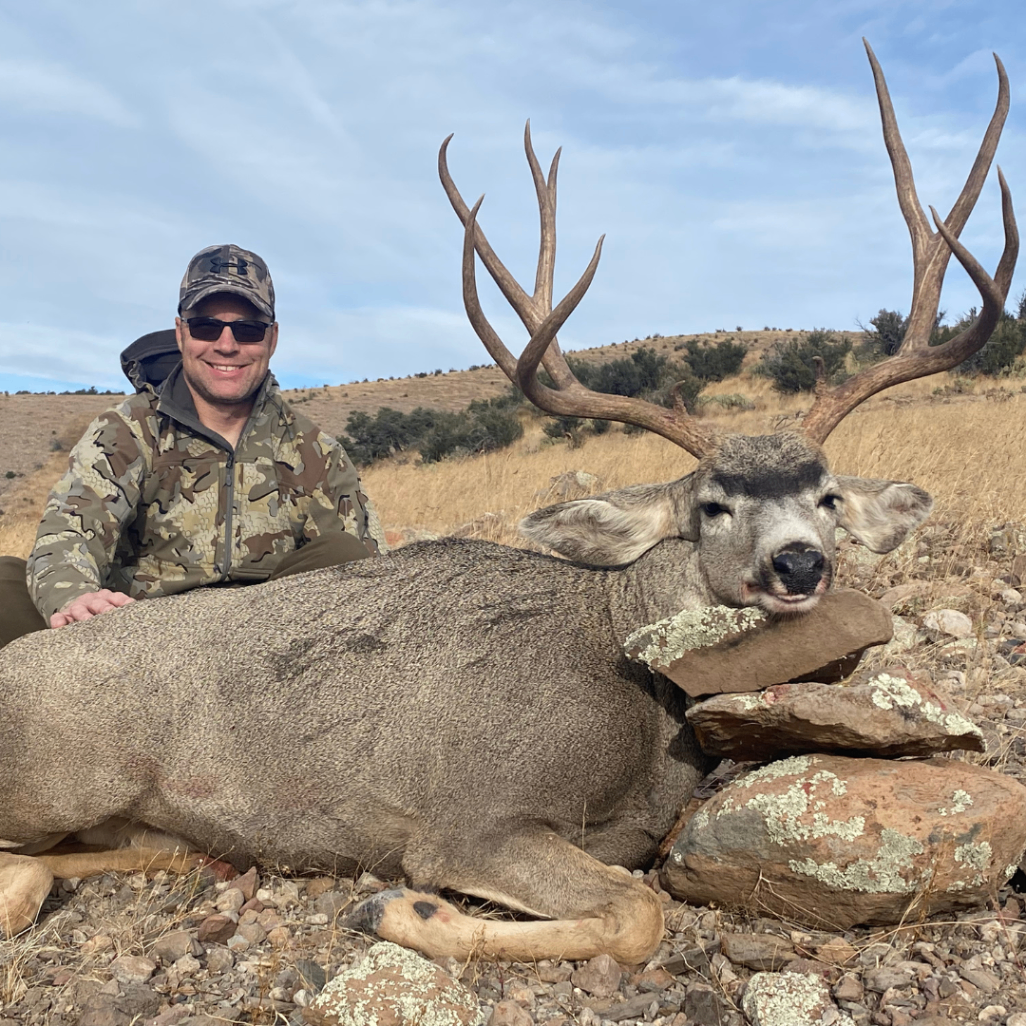 Hunter smiling beside a large mule deer in dry mountain country